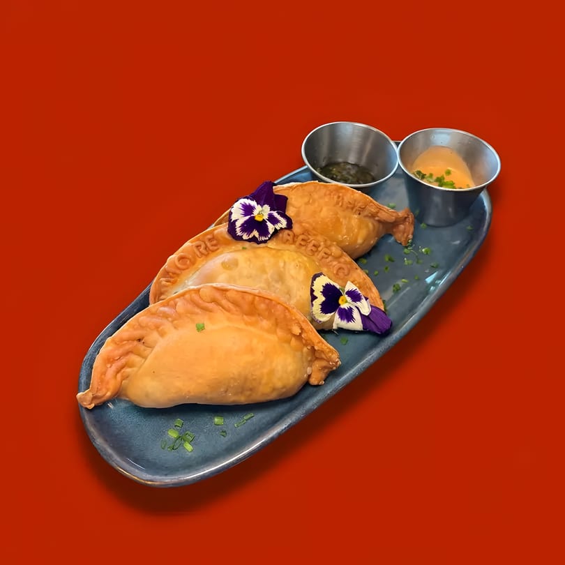 Three golden empanadas garnished with purple edible flowers and two dipping sauces on a blue plate.