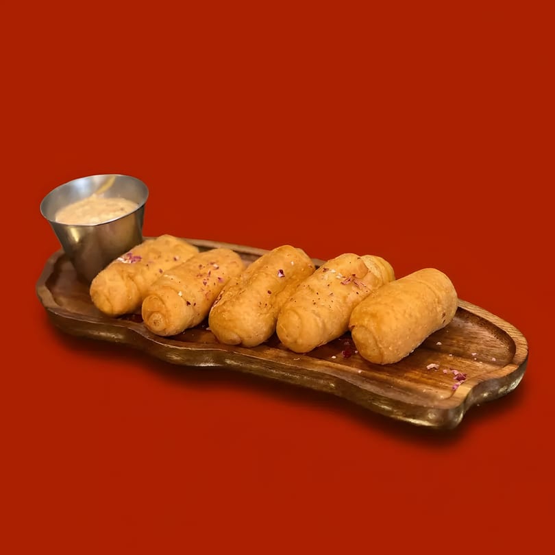 Six golden-brown fried appetizers with a side of dipping sauce on a wooden tray against a red background.